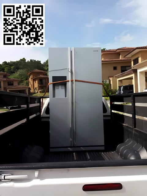 Refrigerador plateado con franja roja cargado en caja de camión frente a casas residenciales y cerca negra