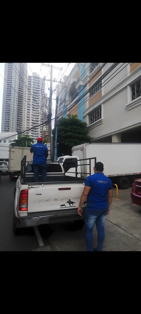 Dos trabajadores en camisas azules en pickup blanco con rack metálico de carga en área residencial urbana